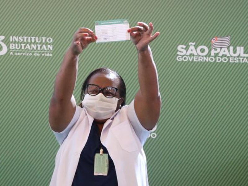 A enfermeira Mônica Calazans segura, com os braços levantados, o comprovante da vacinação, logo após receber a primeira dose da vacina contra a covid. Ela está de óculos,  jaleco branco e máscara